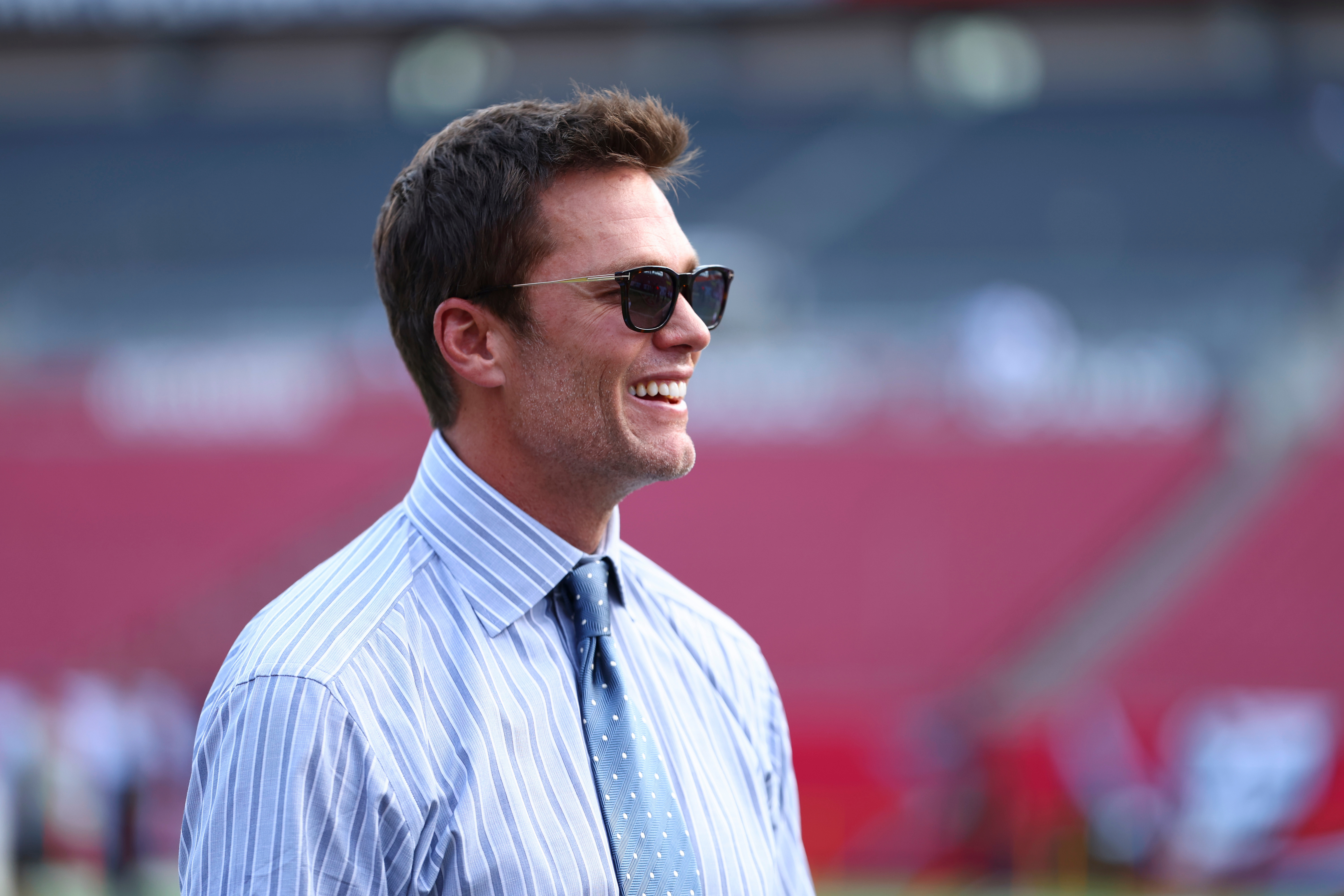 Tom Brady smiling in blue striped dress shirt and sunglasses at NFL stadium