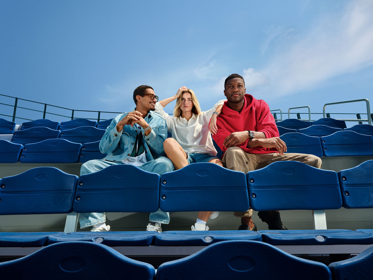 Three young adults wearing Breitling NFL team-edition watches sit casually in blue stadium seats under a clear sky, showcasing the luxury timepieces from the new partnership collection