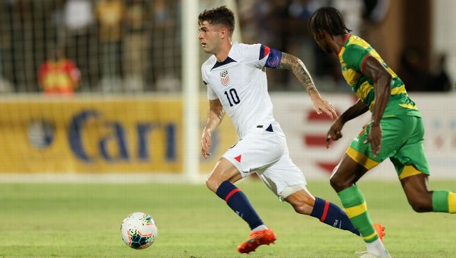 Christian Pulisic number 10 in white USMNT jersey dribbles ball while being pursued by Grenada defender in yellow and green jersey during Nations League match