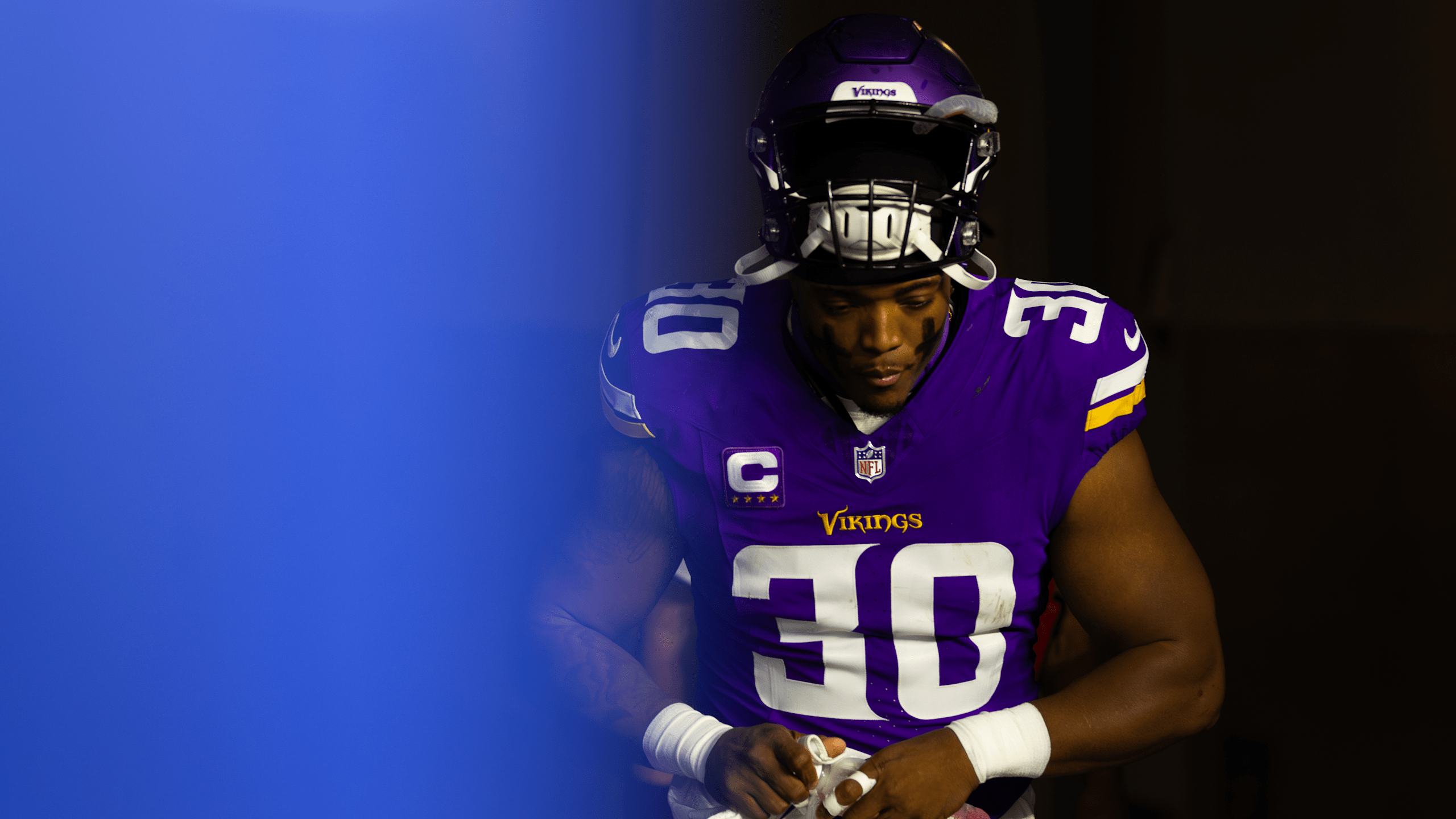 C.J. Ham in Minnesota Vikings purple home uniform with captain's "C" patch, holding a football in dramatic lighting with blue background