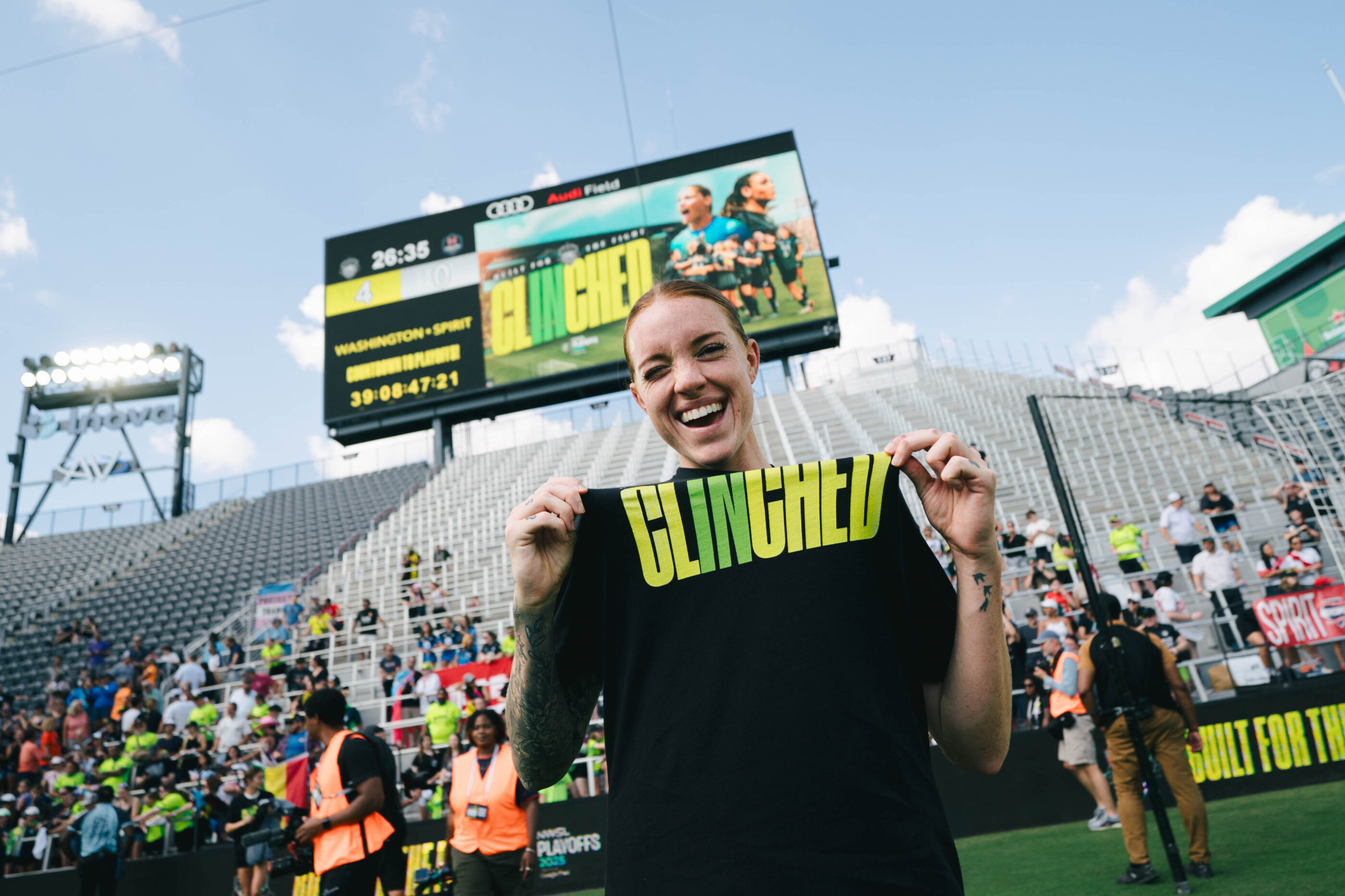 Washington Spirit midfielder Hal Hershfelt smiles while holding up a black "CLINCHED" t-shirt at Audi Field with the stadium scoreboard and crowd visible in the background