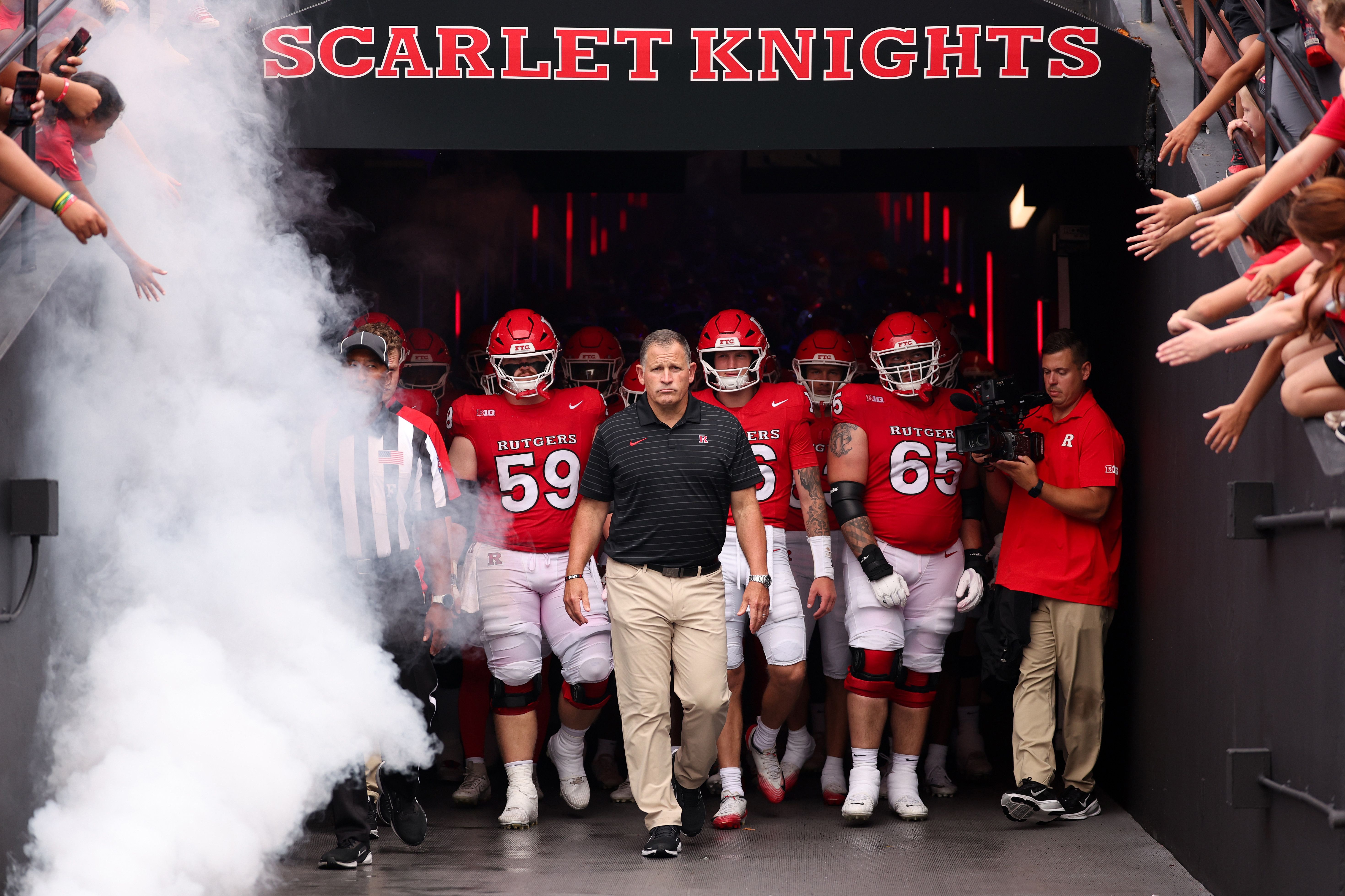 Rutgers Scarlet Knights football team prepares to run onto the field, led by head coach Greg Schiano, with smoke and fans reaching out in the background