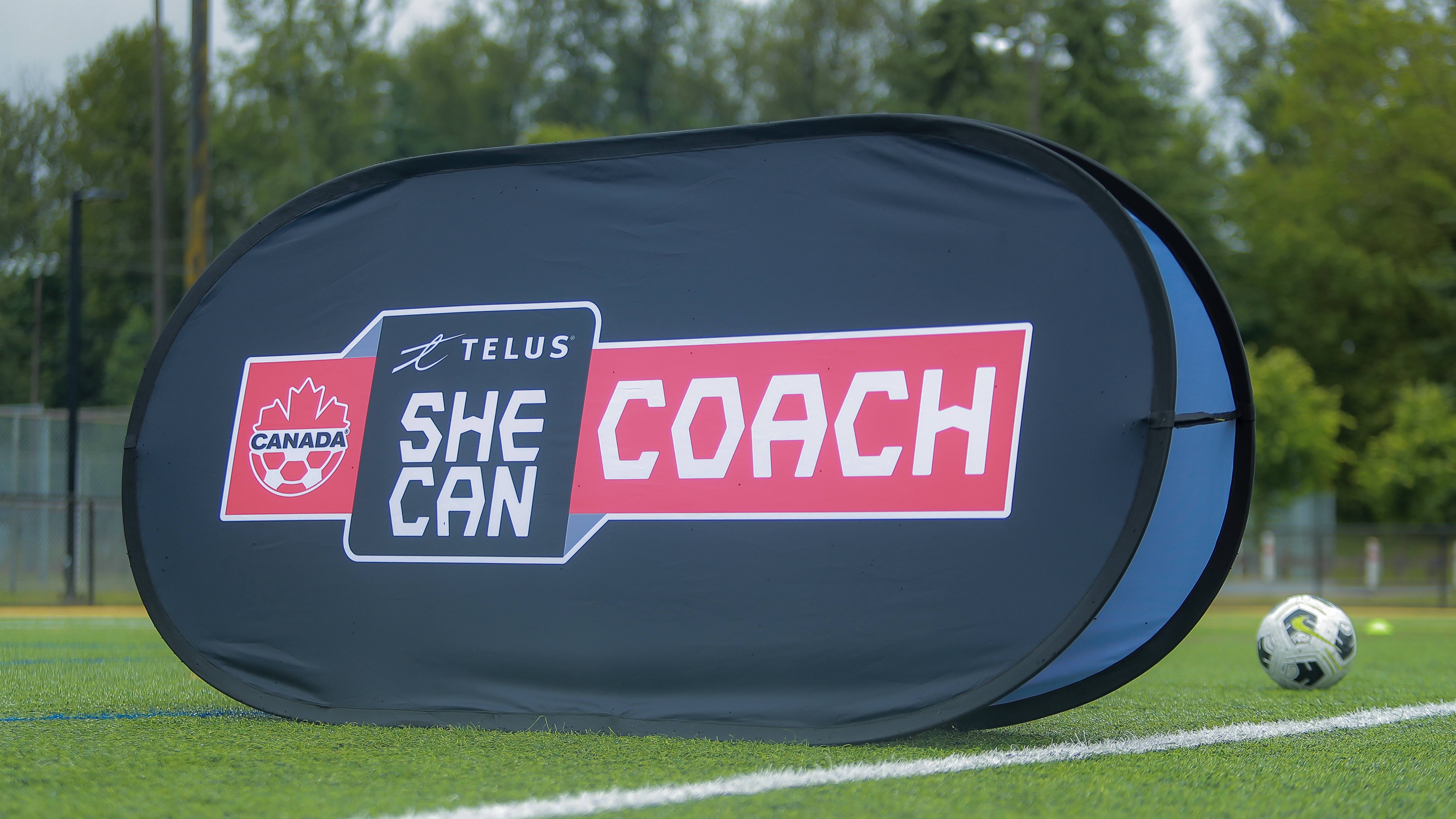 TELUS She CAN Coach branded pop-up banner on a soccer field, featuring Canada Soccer logo, with a soccer ball in the background, symbolizing women's coaching development program