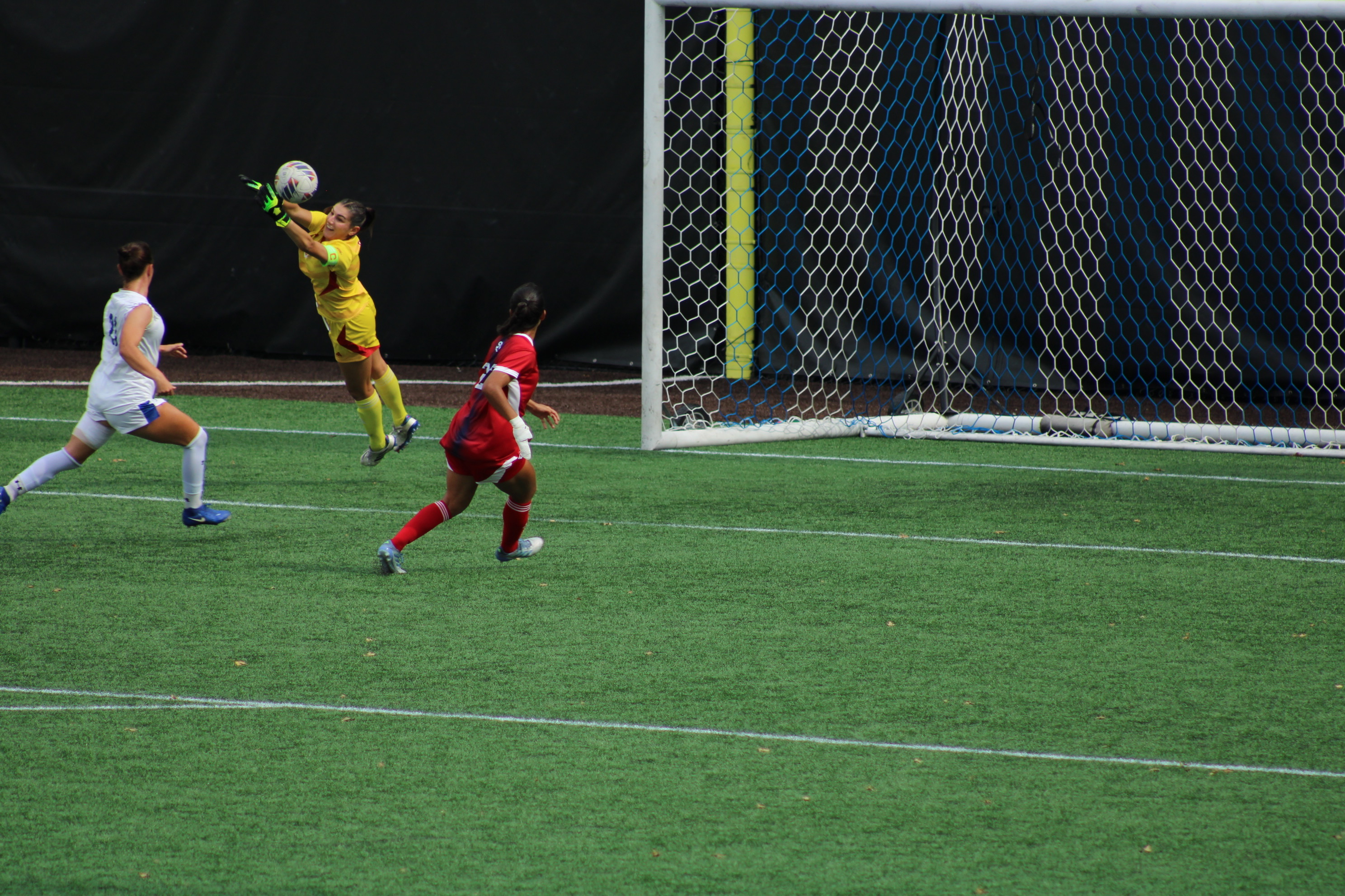 St. John's goalkeeper Kayla Bower in yellow uniform makes a spectacular diving save, leaping high in the air to catch the ball while Seton Hall and St. John's players watch during women's soccer match