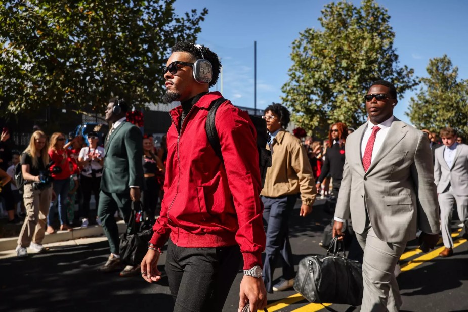 Maryland football players walk into SECU Stadium before their game, dressed in stylish pre-game attire including red team jackets and suits, with fans and media gathered around