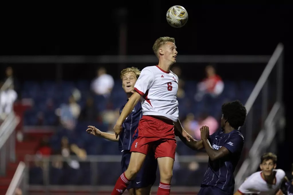 St. John's soccer player Bjorn Nikolajewski, wearing jersey number 6 in white and red, leaps high to win a header against two opposing players in dark blue jerseys during a night match at Belson Stadium