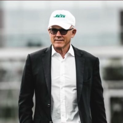 New York Jets owner Woody Johnson walks on the sideline wearing a Jets cap and suit jacket at an NFL game.