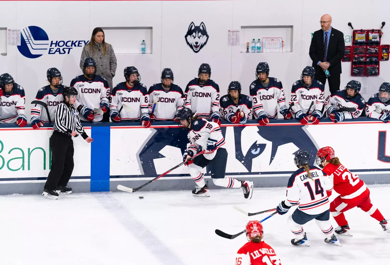 UConn women’s hockey players skate along the boards past a Boston University defender during a Hockey East game, with the UConn bench and coaches in the background.