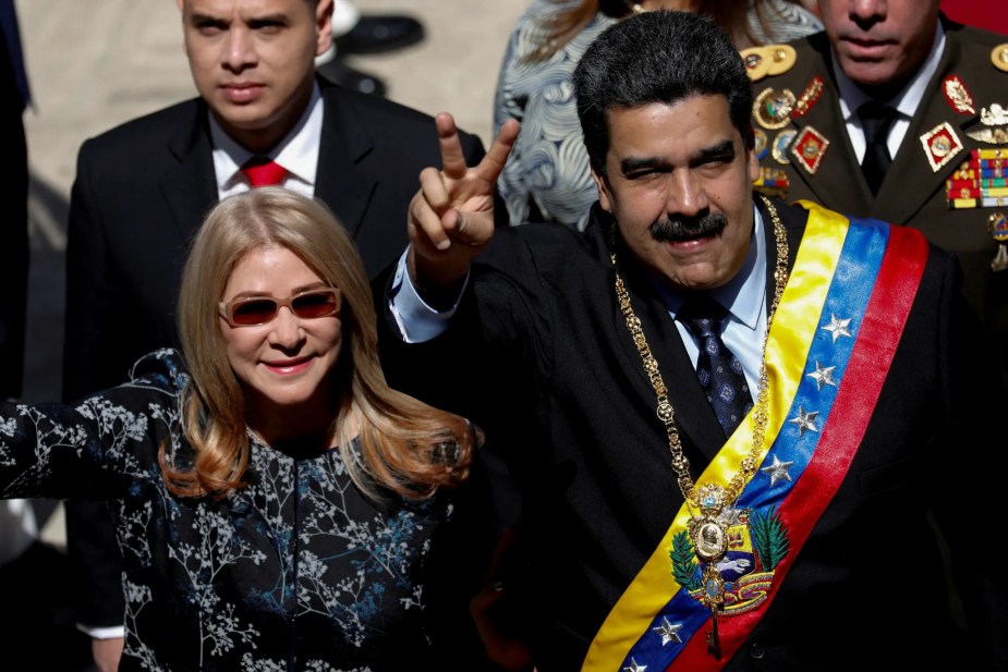 Venezuelan President Nicolás Maduro, wearing a presidential sash and medallion, flashes a peace sign while standing beside his wife, Cilia Flores, during a public event. Military officials are visible in the background.