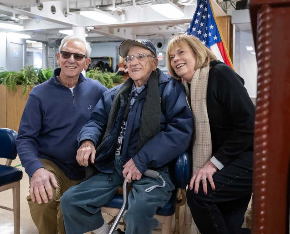 WWII Navy veteran John “Johnny Q” Quinesso Sr. sits smiling between two guests for a photo during his 100th birthday celebration aboard the Battleship New Jersey Museum in Camden, N.J.