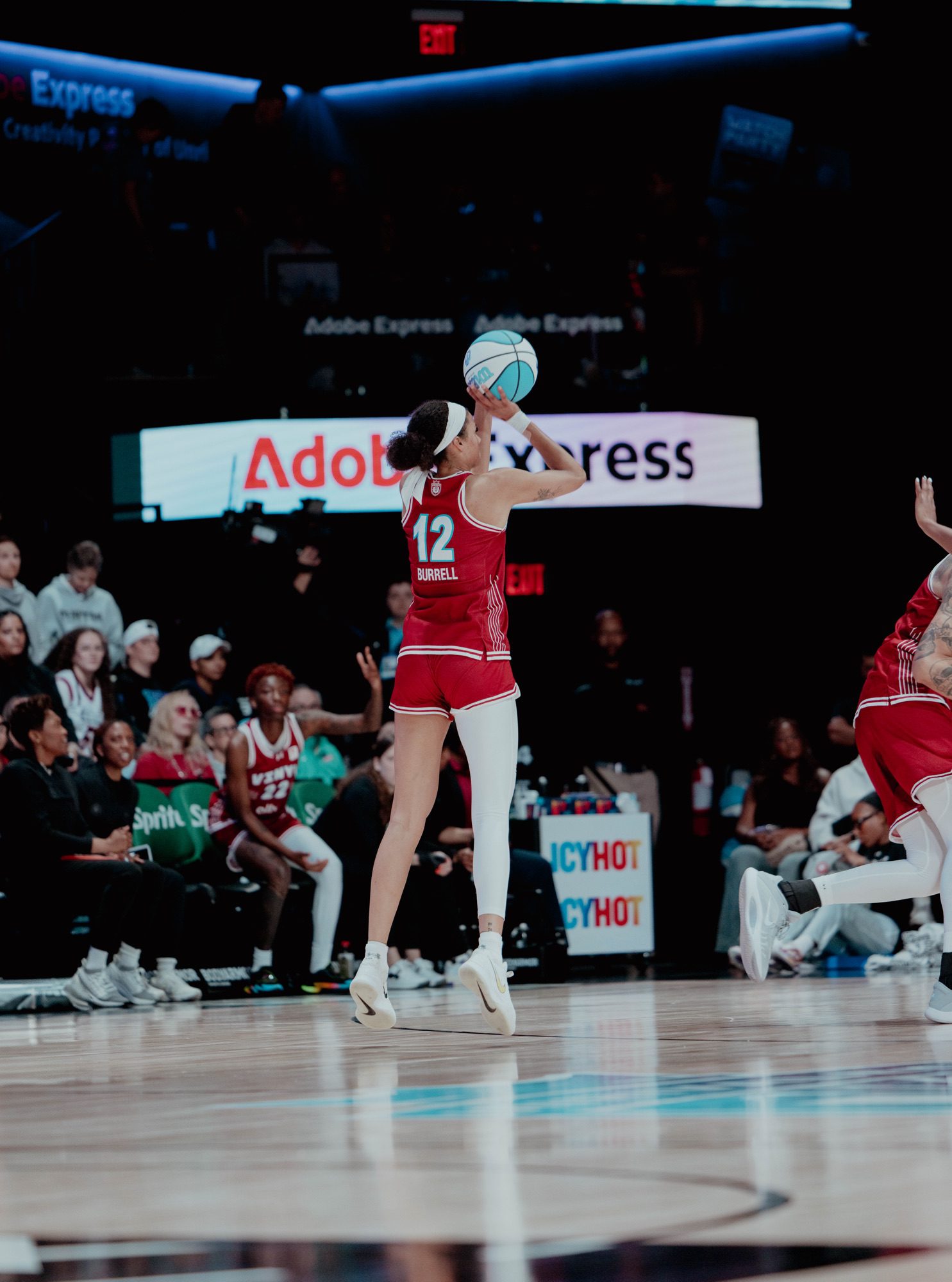 Rae Burrell, wearing a red Vinyl BC jersey with number 12, jumps for a jump shot during an Unrivaled basketball game. She is in mid-air with her arms extended toward a light blue basketball. The background shows a crowded arena with "Adobe Express" and "ICYHOT" signage.